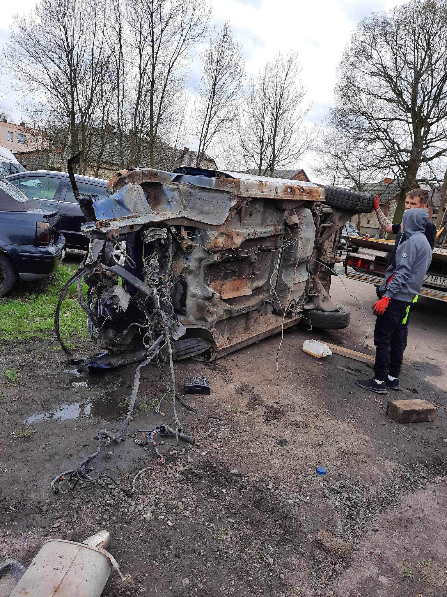 Zniszczony, przewrócony na dach samochód po wypadku, zabezpieczany przez dwóch mężczyzn w odblaskowych elementach i czerwonych rękawicach, widoczna laweta w tle.