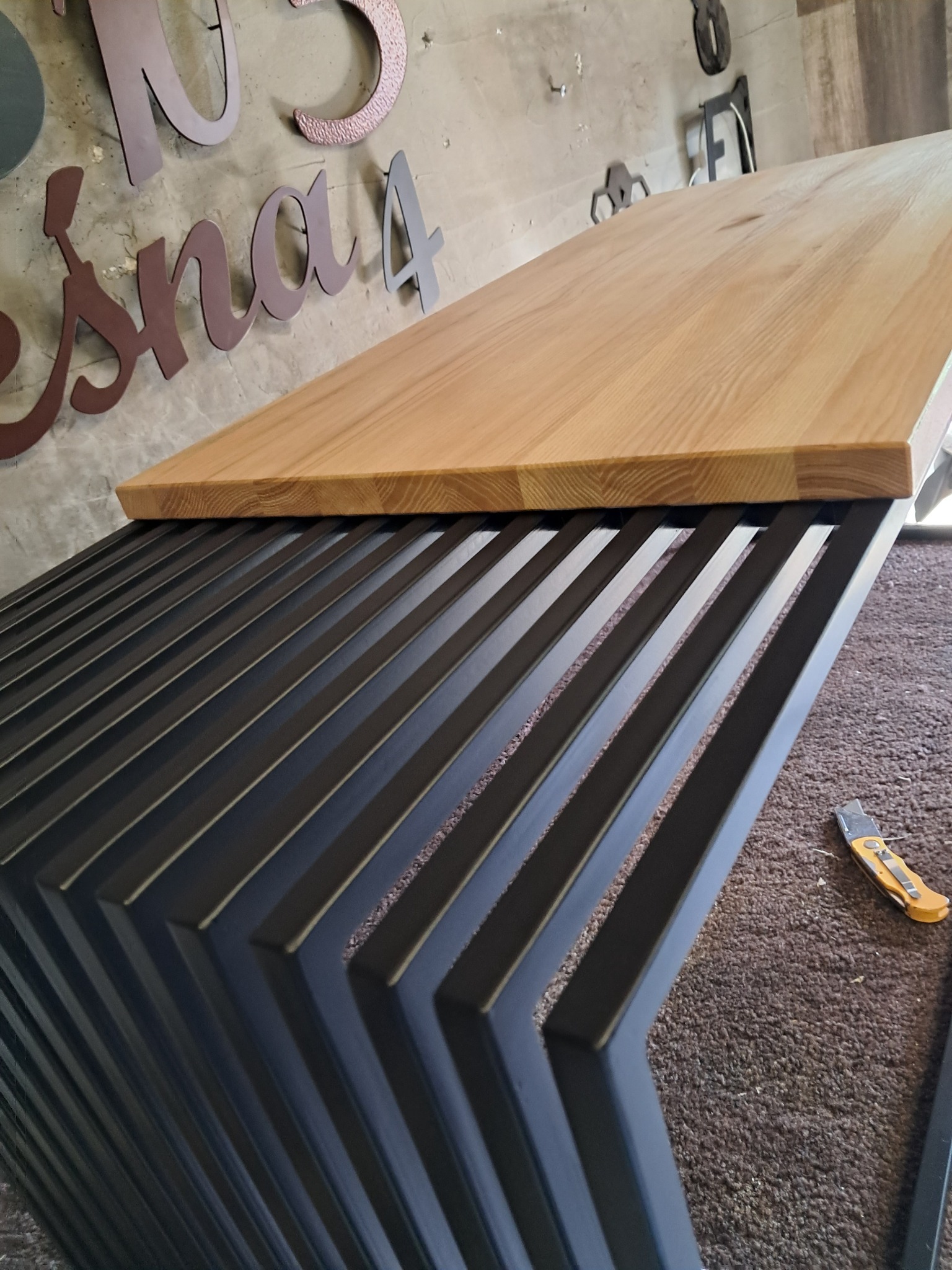 Blat drewniany z jasnego drewna na metalowej, czarnej konstrukcji, widoczny nóż tapicerski na dywanie w tle, dekoracyjne litery na ścianie.