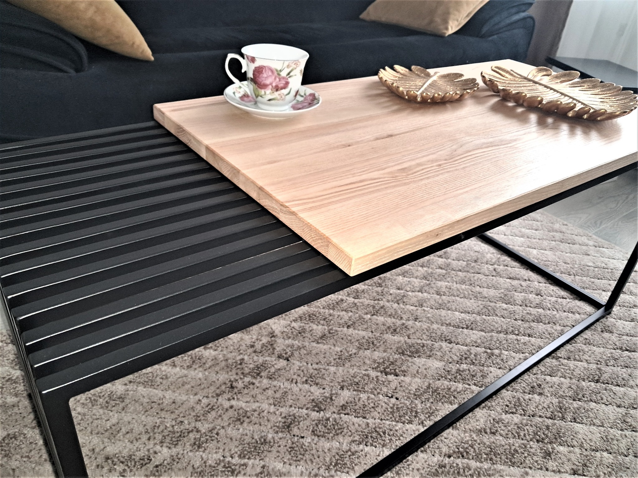 Stolik kawowy z jasnym blatem z drewna i czarną metalową podstawą, na blacie filiżanka z kwiatowym wzorem i dekoracyjne złote liście, w tle sofa z poduszkami.