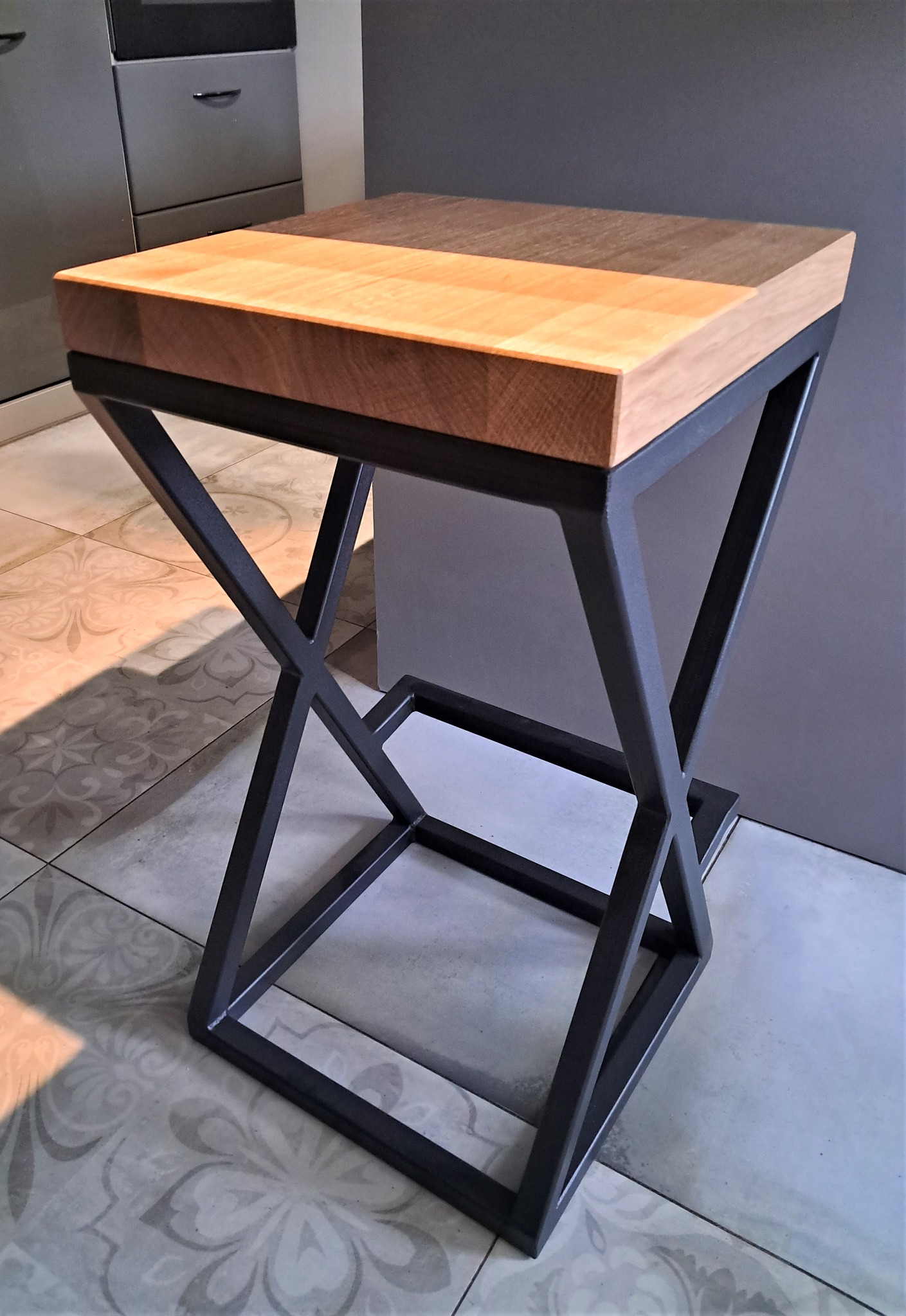 Stolik barowy z blatem z klejonego drewna, o jasnych i ciemnych odcieniach, na metalowej podstawie w kształcie litery X, na tle płytek ceramicznych.