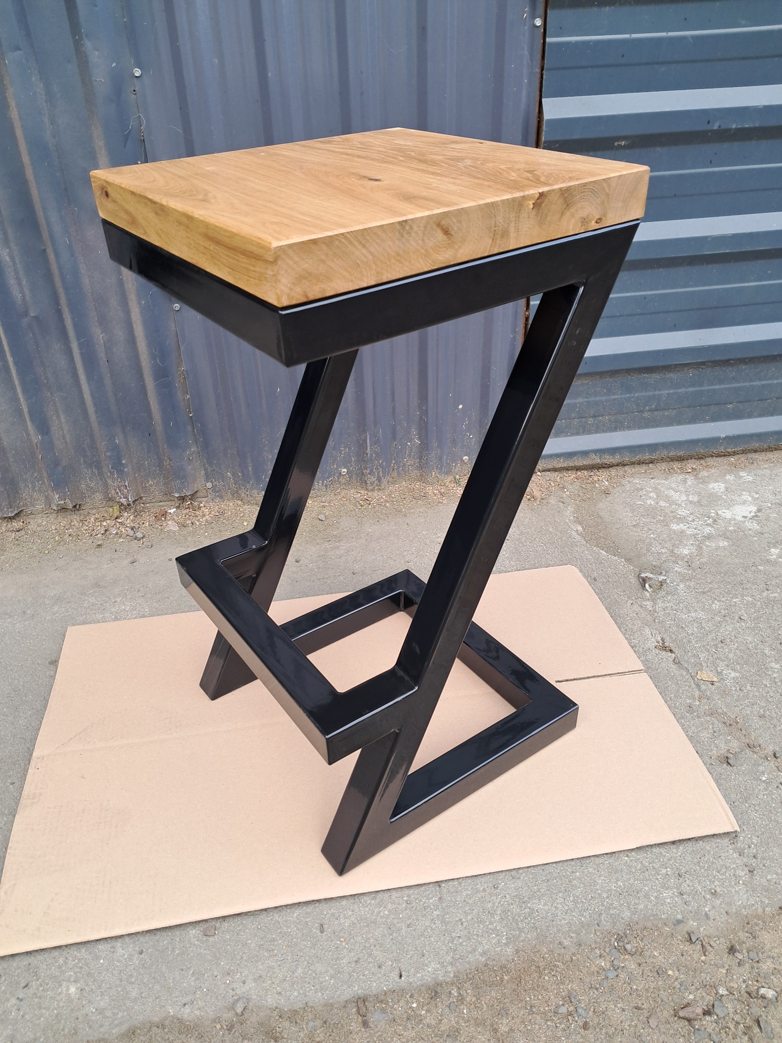 Hoker barowy z blatem z jasnego drewna i czarną, metalową podstawą w kształcie zygzaka, stojący na kartonie na zewnątrz.