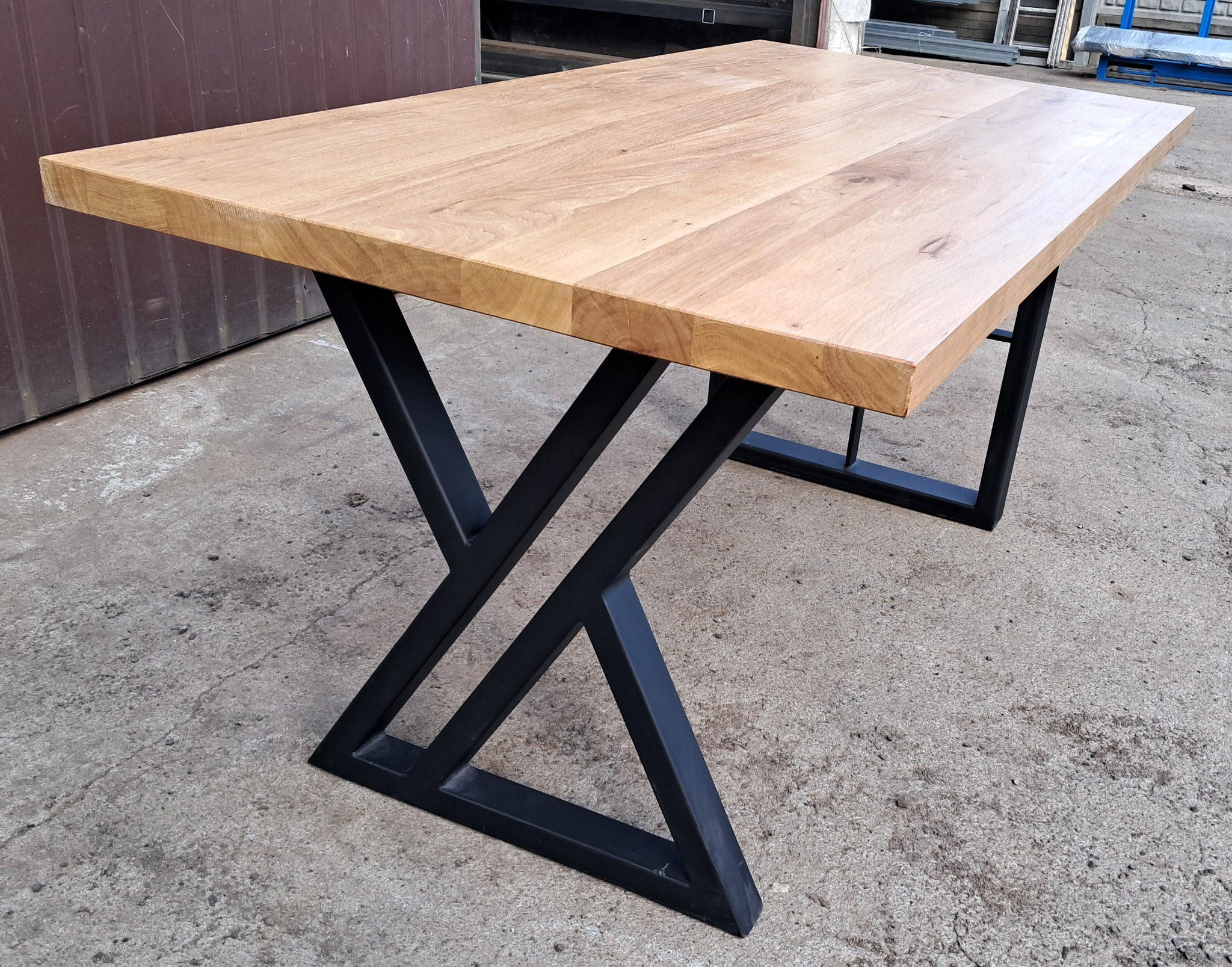 Stół z jasnym blatem z drewna dębowego i czarnymi, geometrycznymi nogami ze stali, na szarym betonowym podłożu.