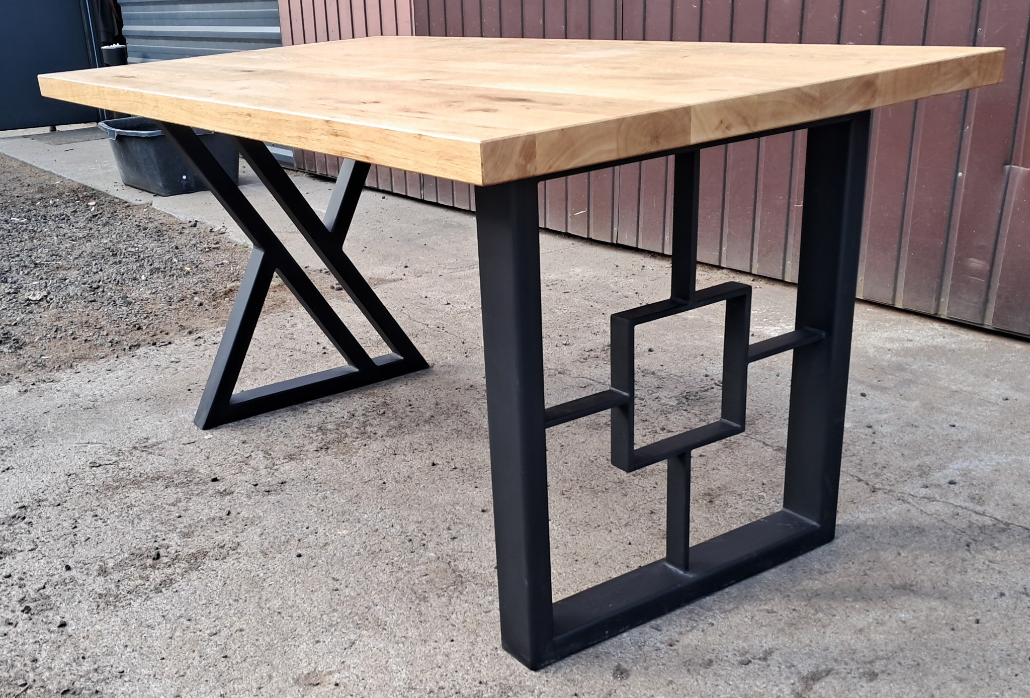 Kwadratowy stół z jasnym blatem drewnianym i czarnymi, metalowymi nogami o geometrycznych wzorach, stojący na betonowym podłożu.