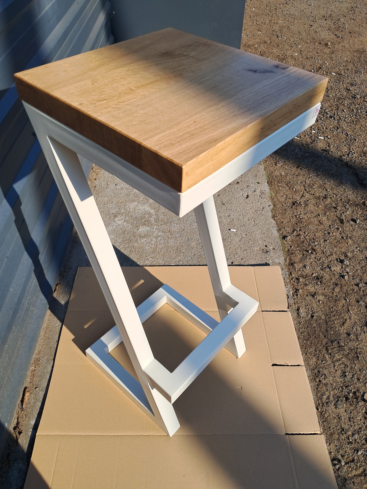 Niewielki, kwadratowy stolik z jasnego drewna na białym, metalowym stelażu o geometrycznym kształcie, sfotografowany w Budzisławiu Kościelnym na tle szarej ziemi i tektury.