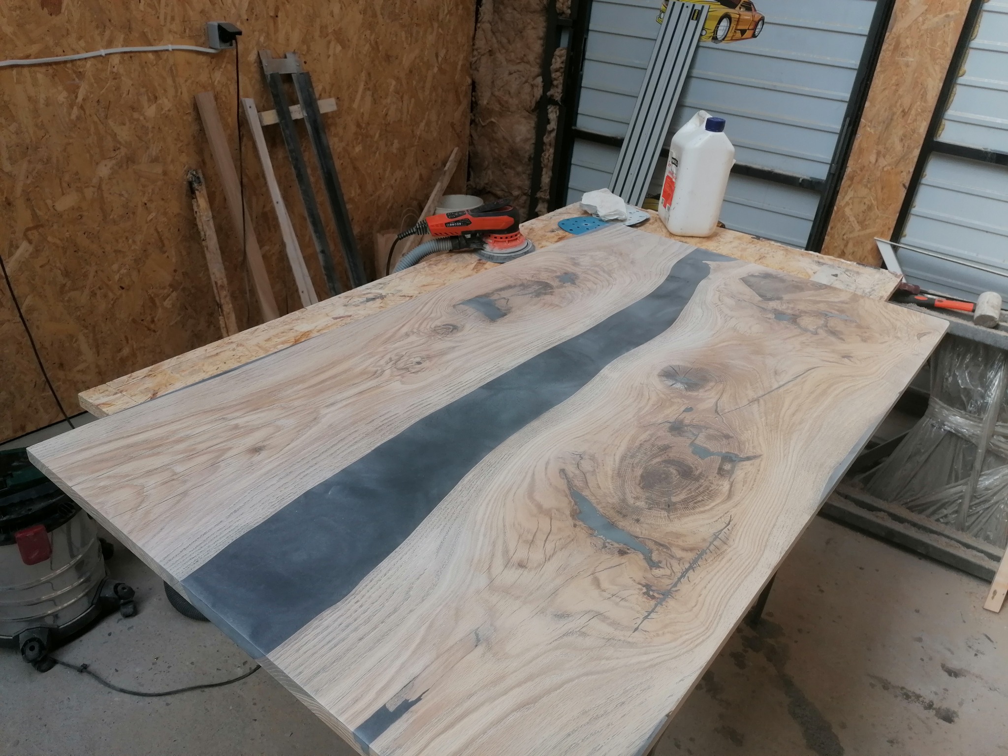 Blat z jasnego drewna z żywicą epoksydową w kolorze szarym, szlifierka elektryczna i akcesoria obok, w warsztacie.