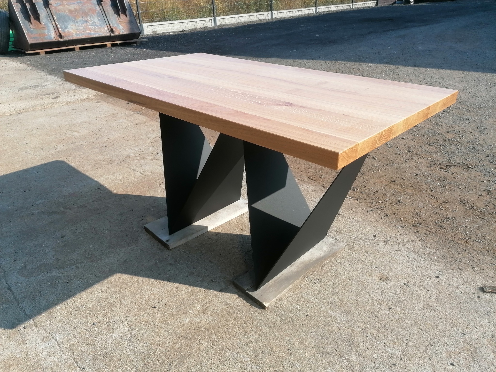 Stół z jasnego drewna z oryginalnymi, geometrycznymi nogami w kolorze czarnym, ustawiony na betonowym podłożu w plenerze.