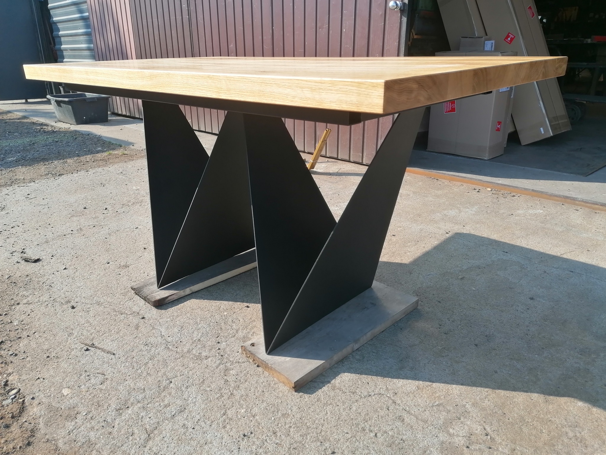 Blat drewniany z jasnego drewna na czarnych, geometrycznych nogach w kształcie litery W, widoczne skrzynie w tle.