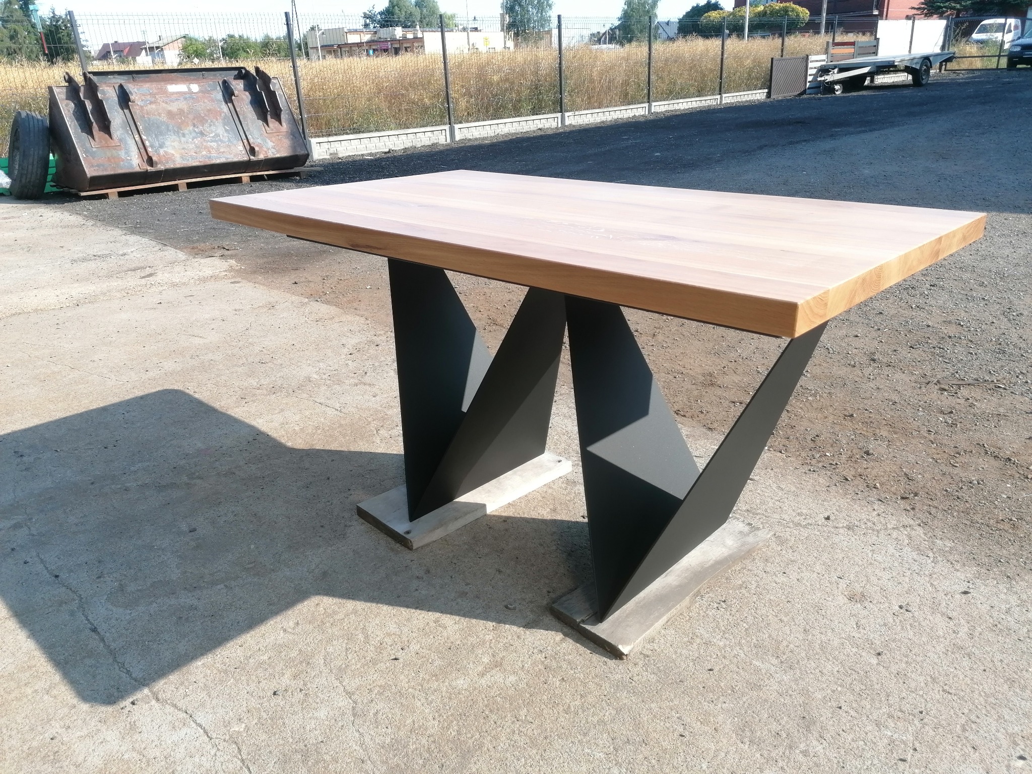 Stół z jasnym blatem i geometrycznymi, czarnymi nogami na betonowych podstawach, na zewnątrz, w tle kontener i trawa.