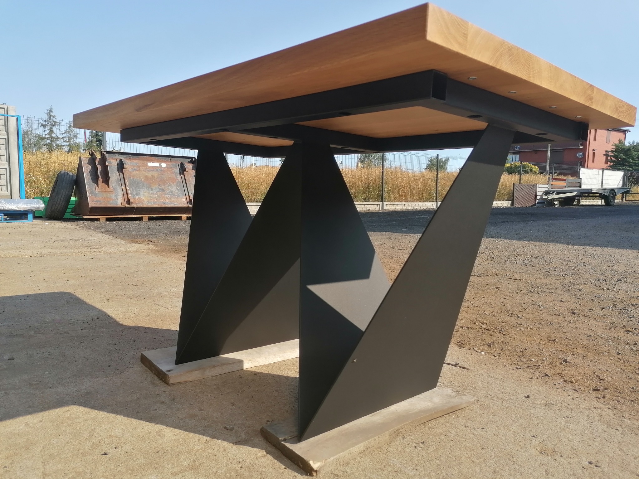 Stół z jasnym blatem drewnianym i geometrycznymi, czarnymi, metalowymi nogami, ustawiony na zewnątrz na betonowej powierzchni. Nogi wsparte na betonowych podkładkach. W tle kontener na śmieci...