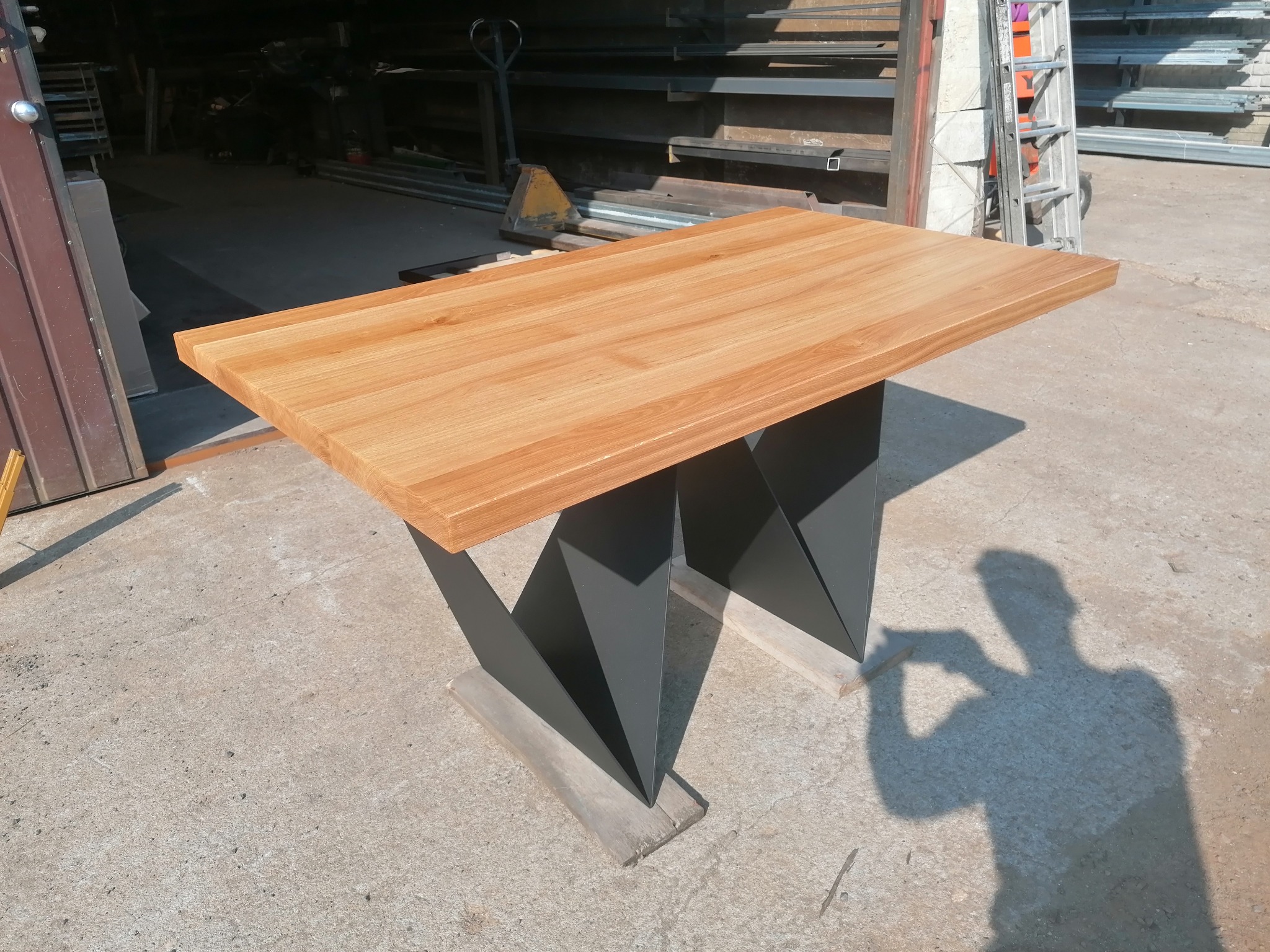 Jasny, dębowy blat na geometrycznej, ciemnoszarej podstawie, ustawiony w warsztacie, z widocznym cieniem fotografa.