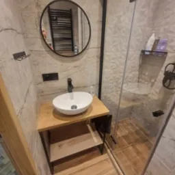 Nowoczesna łazienka z umywalką nablatową na drewnianym blacie, okrągłym lustrem z czarną ramą i kabiną prysznicową z szarymi płytkami imitującymi marmur.