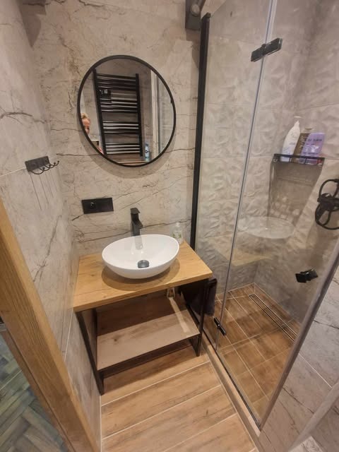 Nowoczesna łazienka z umywalką nablatową na drewnianym blacie, okrągłym lustrem z czarną ramą i kabiną prysznicową z szarymi płytkami imitującymi marmur.