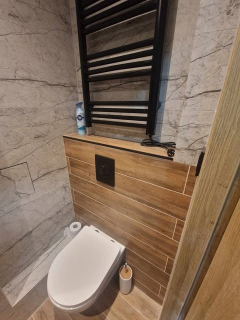 Wykończona łazienka z marmurowymi płytkami, drewnopodobną zabudową, wiszącą toaletą i czarnym grzejnikiem drabinkowym z podłączonym kablem zasilającym.