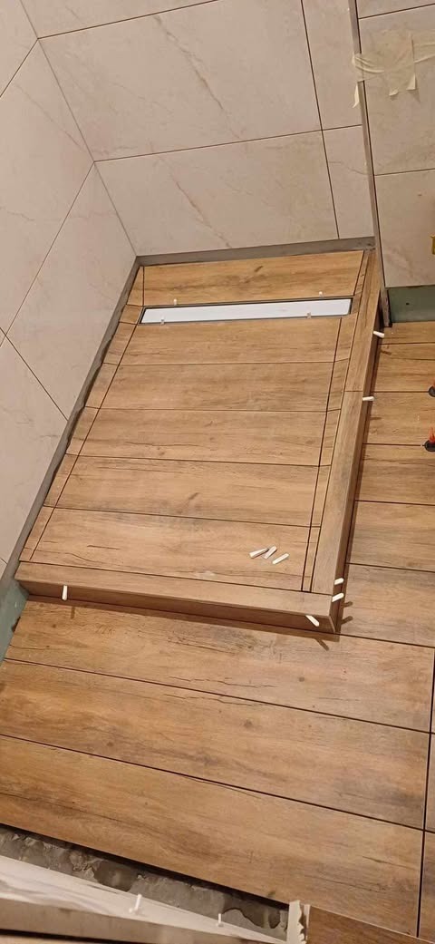 Ukończony brodzik z odpływem liniowym, wyłożony płytkami imitującymi drewno, z widocznymi krzyżykami dystansowymi, na tle wykończonej ściany z jasnych płytek.