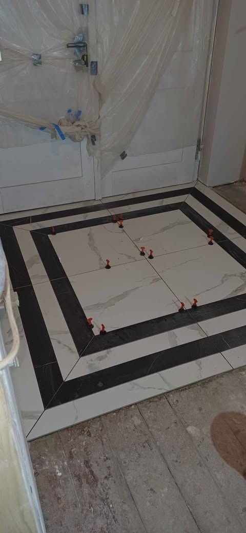 Układanie podłogi z białych i czarnych płytek w kwadratowy wzór, z użyciem czerwonych klipsów poziomujących, drzwi zabezpieczone folią ochronną.