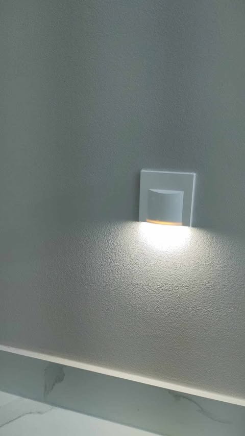 Ściana z fakturą pomalowana na szaro, oświetlona przez nowoczesny kinkiet LED z ciepłym światłem.