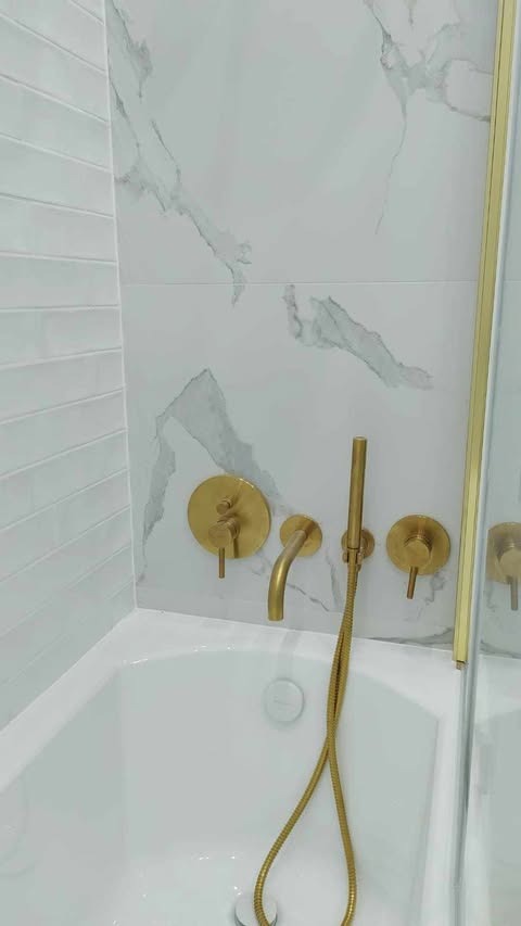 Biała wanna z baterią w kolorze szczotkowanego złota i deszczownicą na tle płytek imitujących marmur i białych płytek ułożonych w cegiełkę.