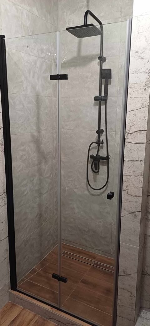 Kabina prysznicowa z czarnymi profilami i deszczownicą, wykończona płytkami imitującymi marmur, z drewnopodobną podłogą i odpływem liniowym.