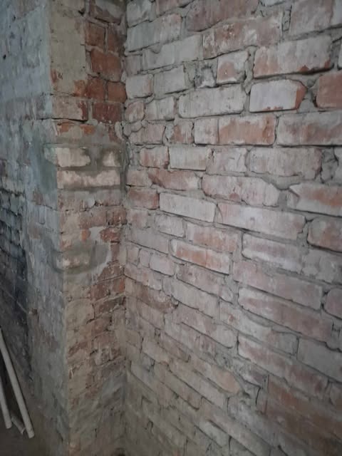 Fragment narożnika pomieszczenia z surową ścianą z czerwonej cegły, częściowo pokrytej szarym tynkiem, widoczne białe rury przy ścianie.
