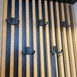 Małgorzata Kosek Zakład Produkcyjno-Usługowo-Handlowy Kosmebels - Panel ścienny z drewnianych listew w kolorze jasnego dębu, przeplatanych czarnymi pasami, z przymocowanymi czarnymi, metalowymi wieszakami na ubrania; na jednym z wieszaków wisi szary łyżka do butów.