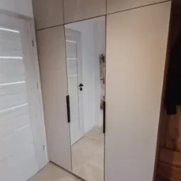 Jasnoszara szafa wnękowa z lustrzanymi drzwiami, odbijająca wnętrze pokoju z widocznymi drzwiami, zamontowana w mieszkaniu w Izdebniku. Minimalistyczny design z długimi, czarnymi uchwytami.