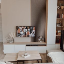 Małgorzata Kosek Zakład Produkcyjno-Usługowo-Handlowy Kosmebels - Jasny salon z zabudową meblową: szafki RTV z białymi frontami i drewnianym blatem, pionowe drewniane lamele dekoracyjne, regał z książkami, abstrakcyjny obraz nad szafkami, kawa na stoliku kawowym.