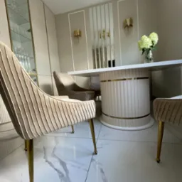 Elegancka jadalnia z okrągłym stołem na nodze z pionowymi żłobieniami i aksamitnymi krzesłami na złotych nóżkach, uzupełniona szklaną witryną i dekoracyjnymi kinkietami.