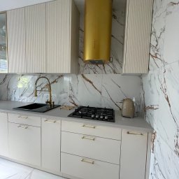 Małgorzata Kosek Zakład Produkcyjno-Usługowo-Handlowy Kosmebels - Nowoczesna kuchnia z beżowymi frontami szafek z delikatnym frezowaniem, złotymi uchwytami i złotym okapem na tle marmurowych płytek.