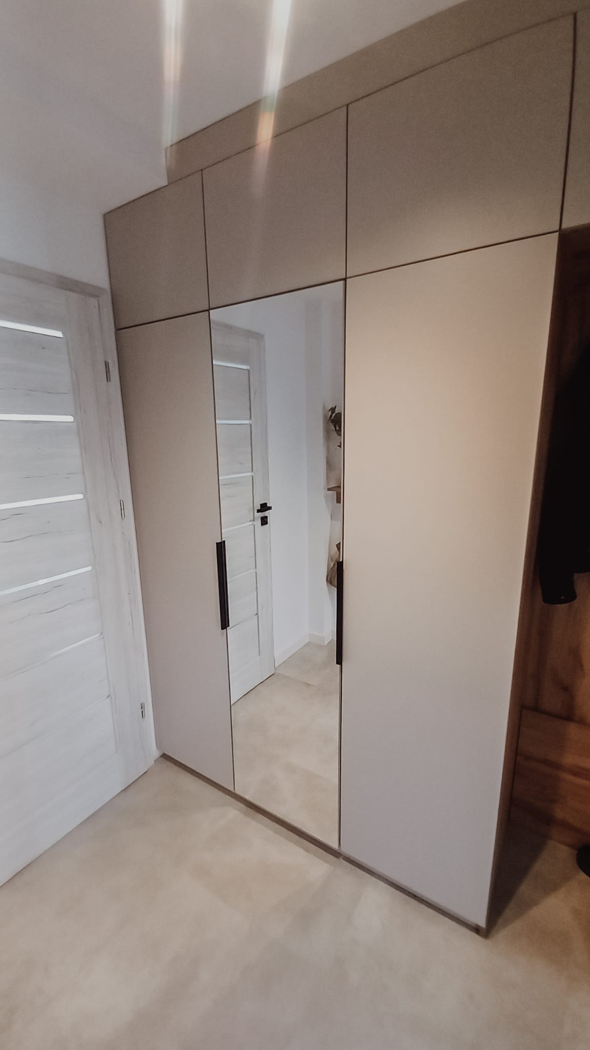 Jasnoszara szafa wnękowa z lustrzanymi drzwiami, odbijająca wnętrze pokoju z widocznymi drzwiami, zamontowana w mieszkaniu w Izdebniku. Minimalistyczny design z długimi, czarnymi uchwytami.
