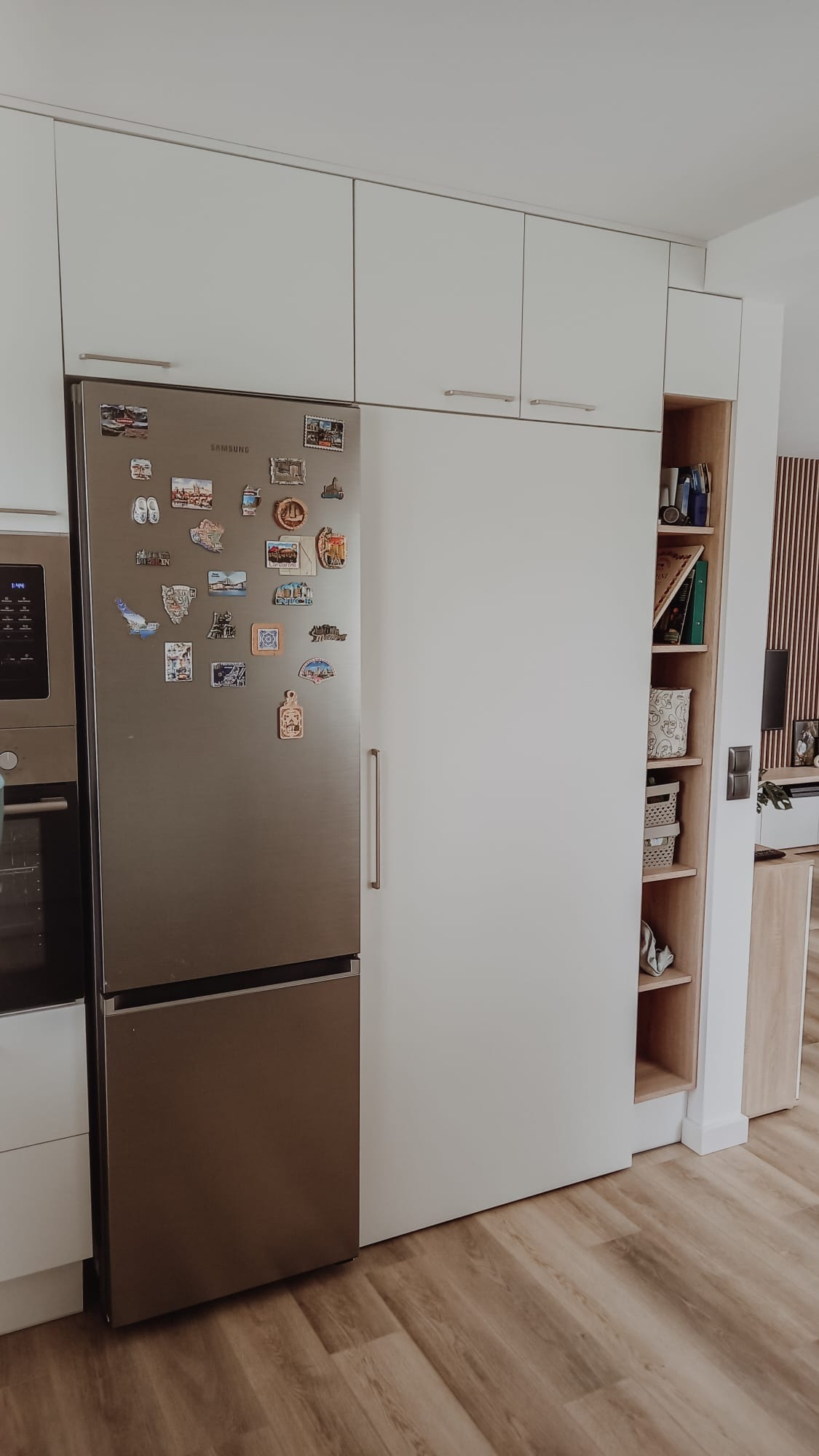 Biała zabudowa kuchenna z lodówką obłożoną magnesami z podróży i otwartym regałem z dekoracjami.