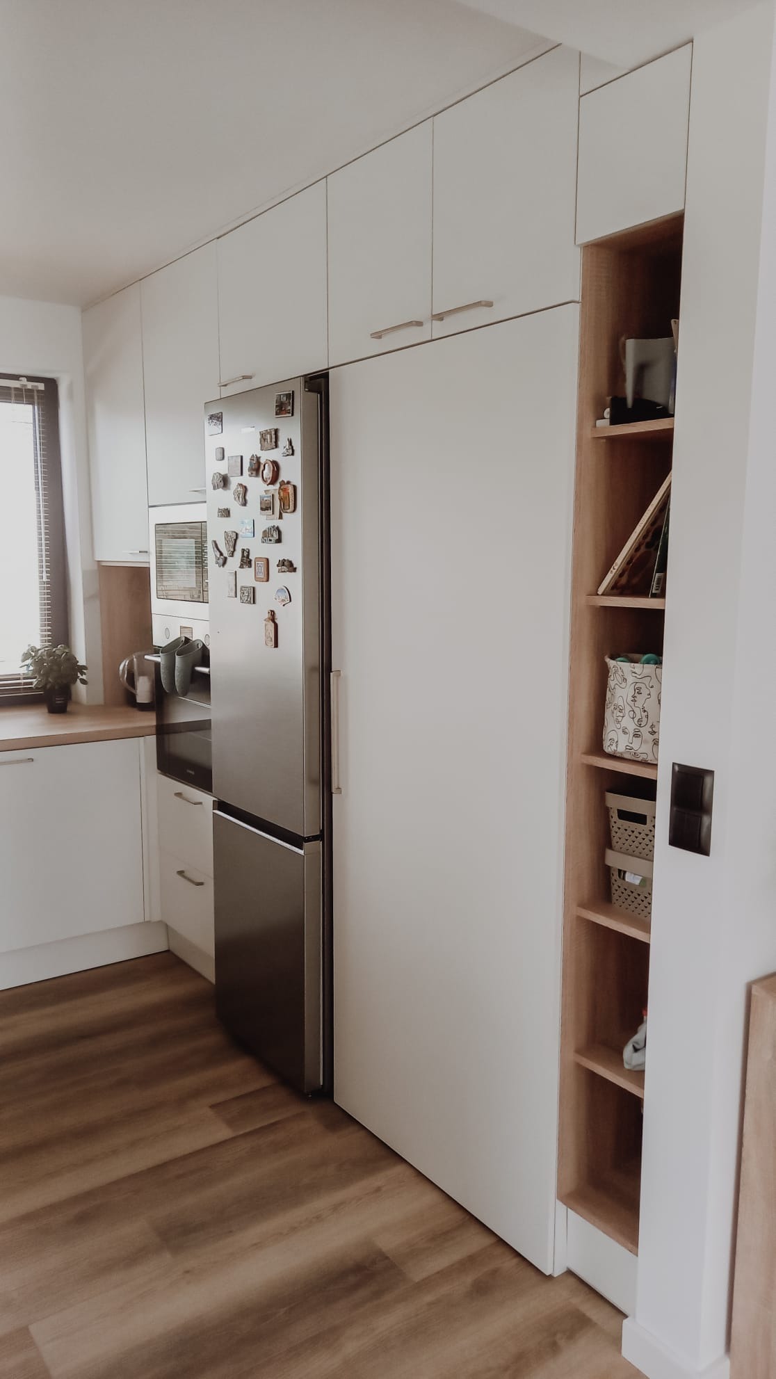 Nowoczesna kuchnia z białymi szafkami na wymiar, srebrną lodówką z magnesami, drewnianym blatem i otwartym regałem z półkami z jasnego drewna.