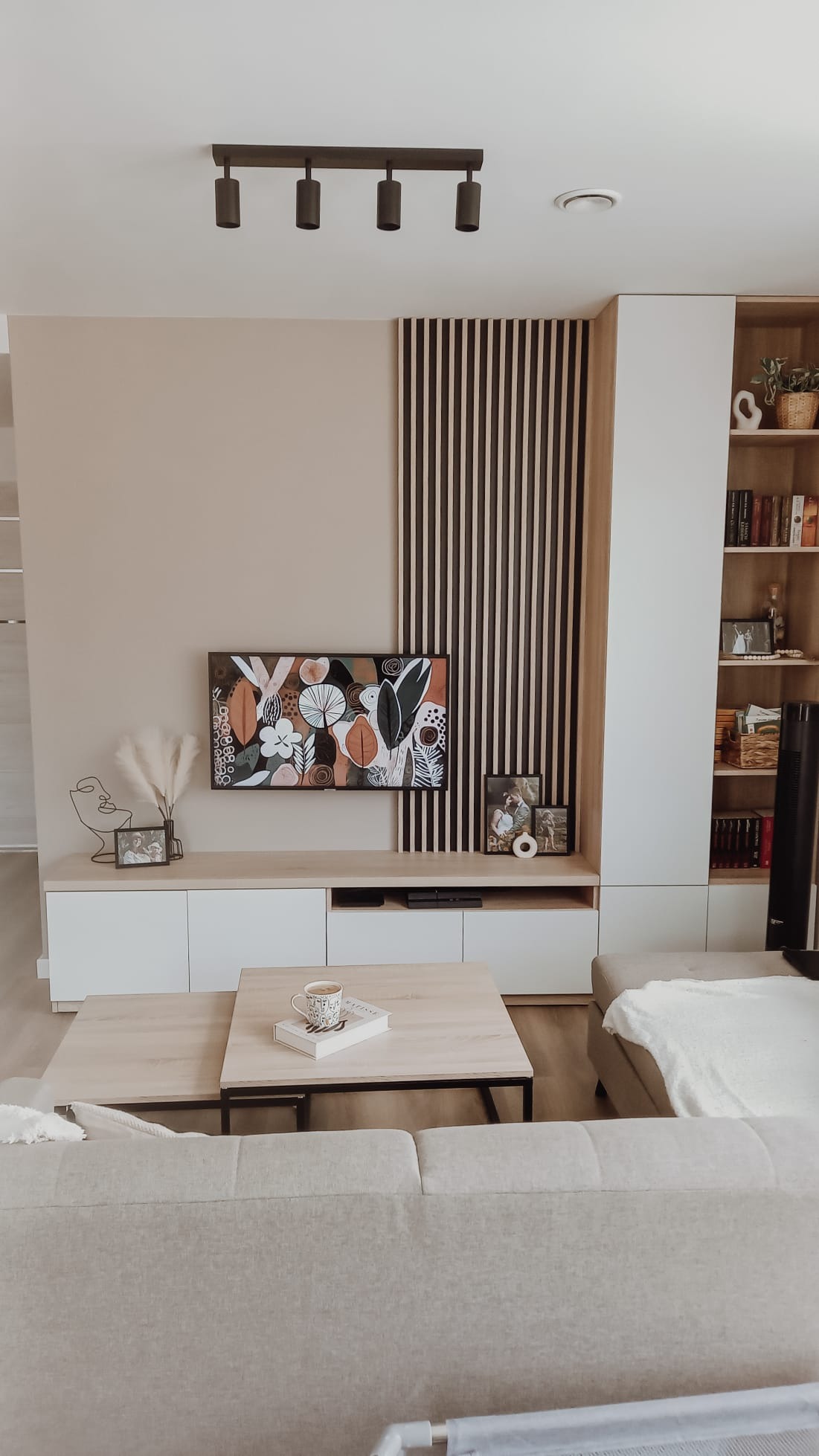 Jasny salon z zabudową meblową: szafki RTV z białymi frontami i drewnianym blatem, pionowe drewniane lamele dekoracyjne, regał z książkami, abstrakcyjny obraz nad szafkami, kawa na stoliku kawowym.