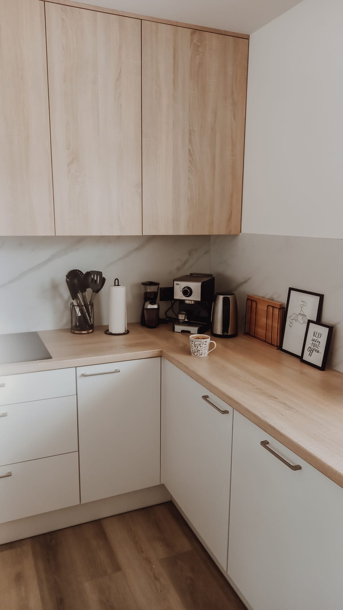 Jasna kuchnia z drewnianym blatem, białymi szafkami i sprzętami AGD, uzupełniona dekoracjami, takimi jak kubek z wzorem i ramki z napisami.