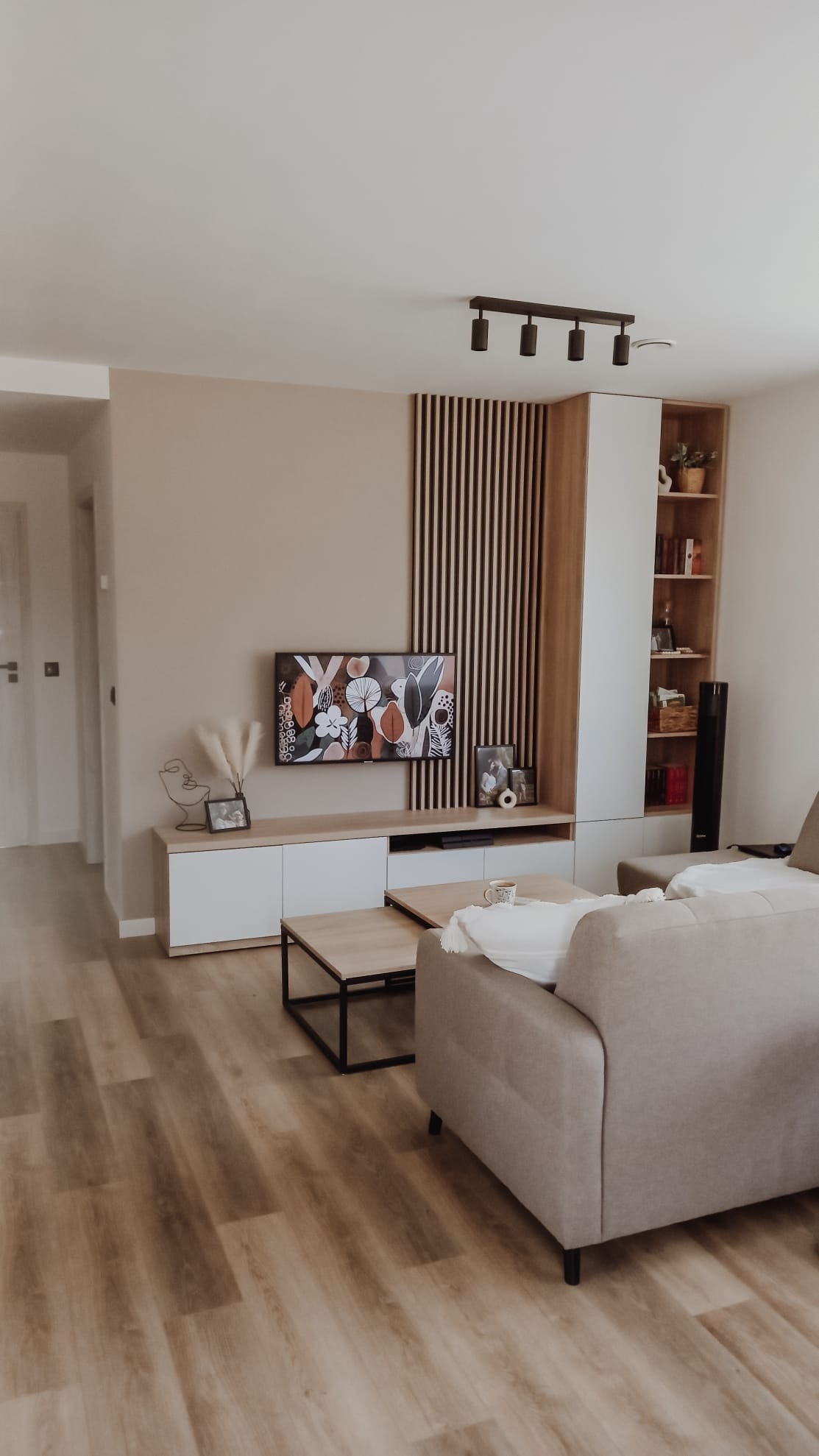 Jasny salon z drewnianą podłogą, beżową ścianą, telewizorem, szafką RTV z białymi frontami, drewnianymi lamelami dekoracyjnymi na ścianie, regałem z półkami i beżową sofą.