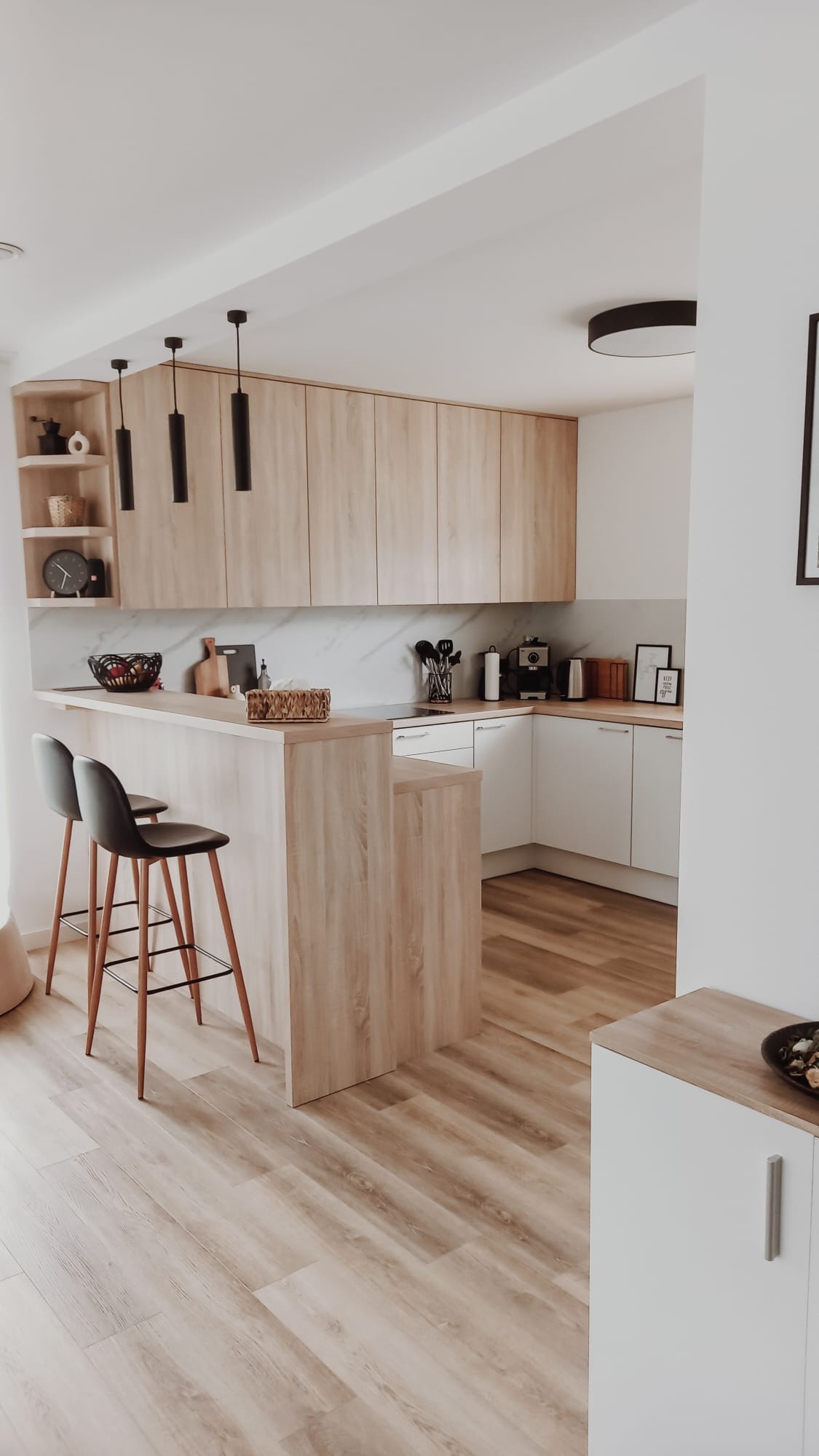 Jasna kuchnia z drewnianymi frontami szafek górnych i barkiem oddzielającym przestrzeń, dwa czarne hokery przy barku, drewniana podłoga.