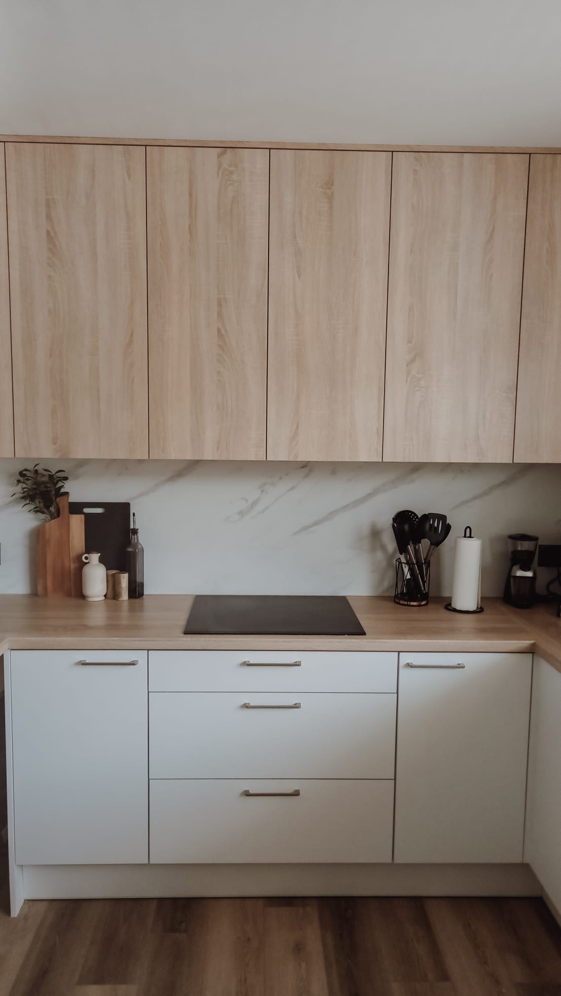 Jasna kuchnia z drewnianymi szafkami górnymi i białymi dolnymi, blatem w kolorze drewna i indukcyjną płytą grzewczą, minimalistyczny wystrój z akcesoriami kuchennymi.