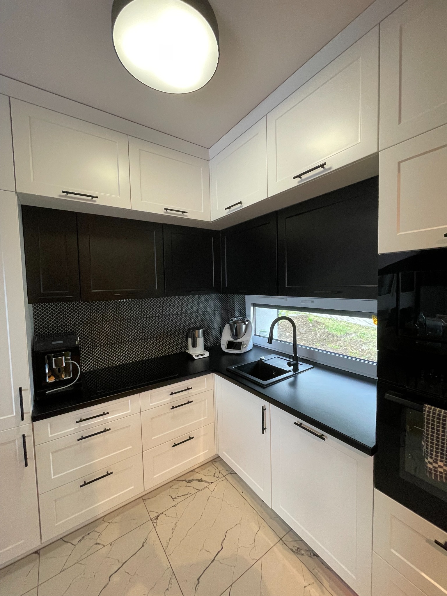 Nowoczesna kuchnia w bieli i czerni z czarnym blatem, mozaiką nad blatem i marmurową podłogą, oświetlona okrągłą lampą sufitową.