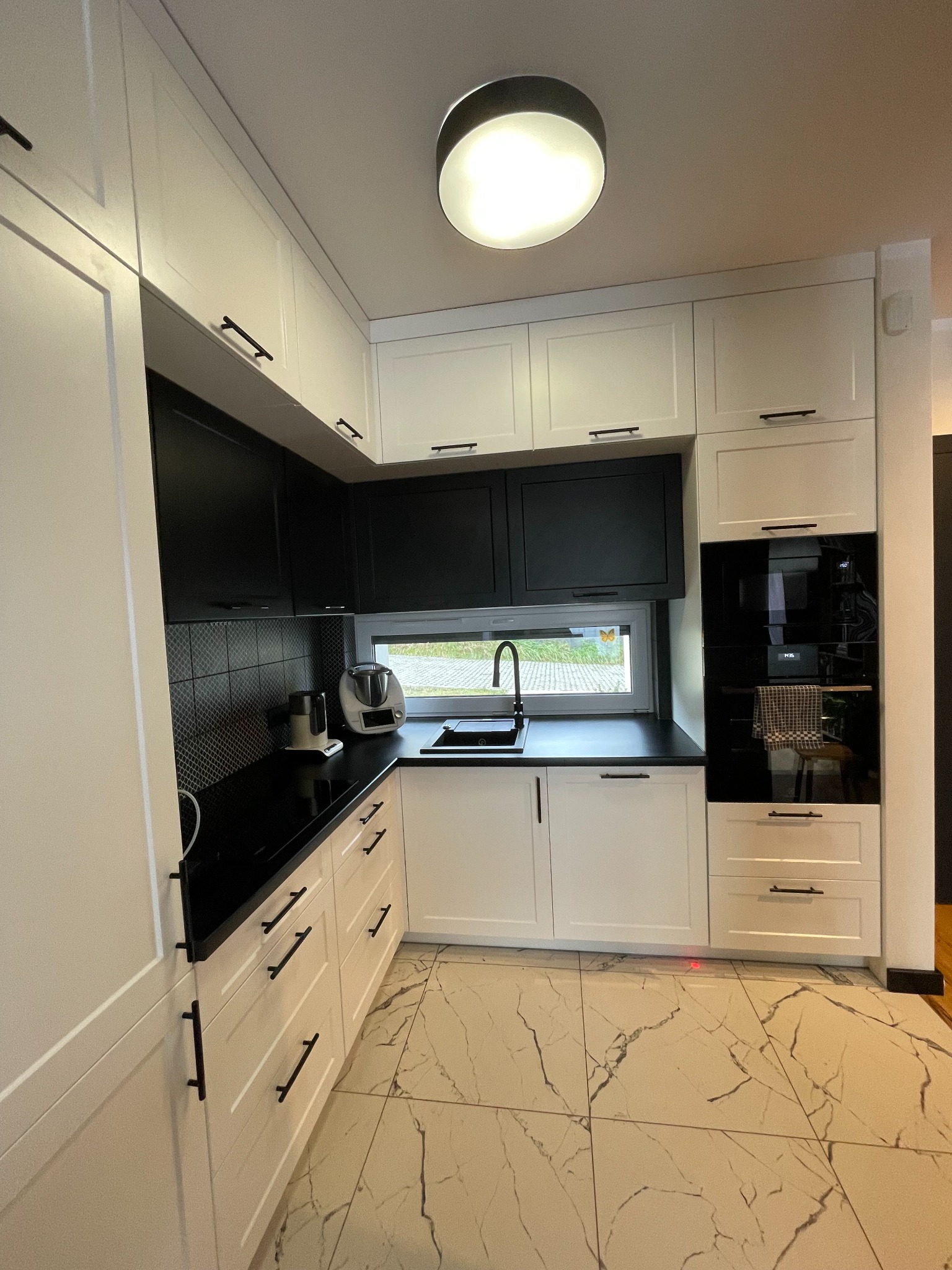 Nowoczesna kuchnia z białymi szafkami dolnymi i górnymi, czarnymi szafkami wiszącymi, czarnym blatem i czarną armaturą, uzupełniona o marmurowe płytki podłogowe.