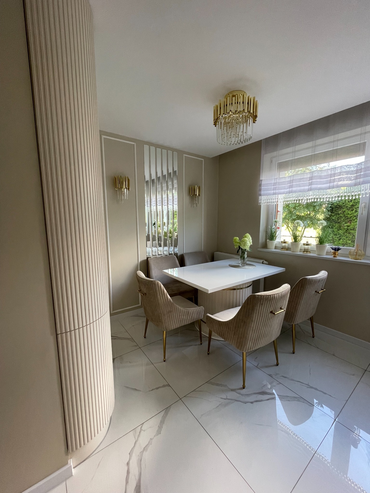 Elegancka jadalnia w beżach: stół z białym blatem, tapicerowane krzesła na złotych nóżkach, dekoracyjny lamel na ścianie, złoty żyrandol i lustrzane panele na ścianie.