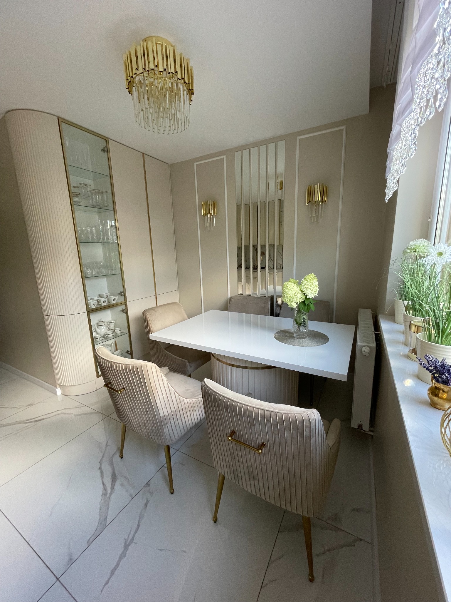 Elegancka jadalnia w odcieniach beżu i bieli: przeszklona witryna z zastawą, stół z białym blatem i welurowe krzesła na złotych nóżkach, doświetlona złotym żyrandolem i kinkietami.