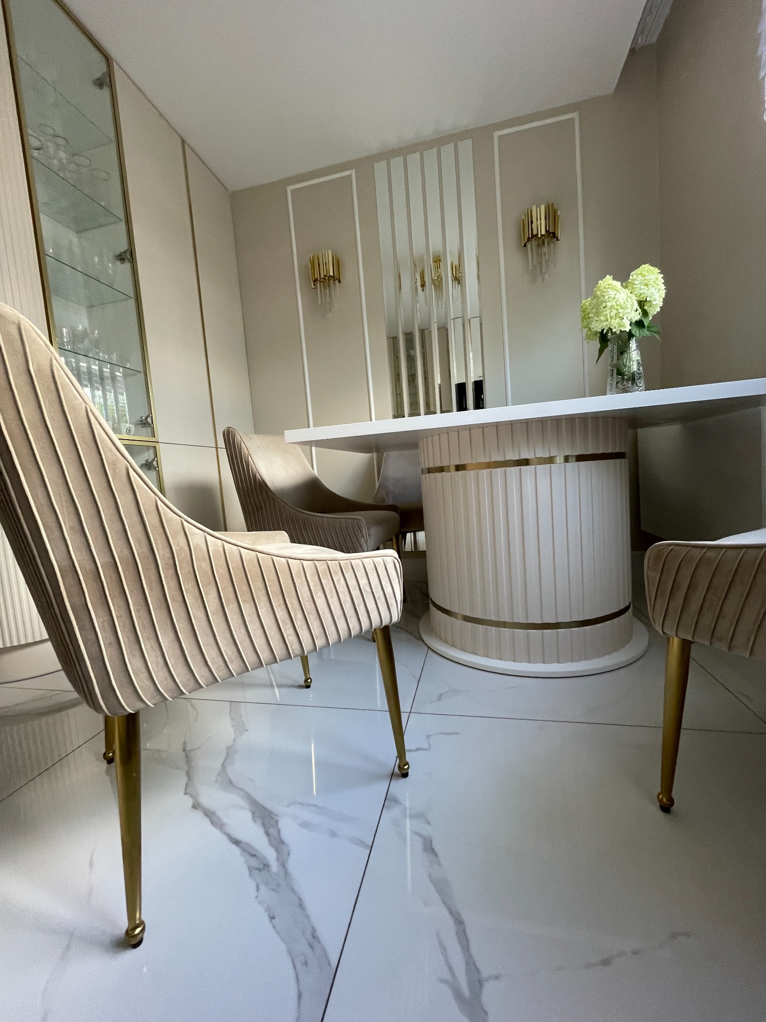 Elegancka jadalnia z okrągłym stołem na nodze z pionowymi żłobieniami i aksamitnymi krzesłami na złotych nóżkach, uzupełniona szklaną witryną i dekoracyjnymi kinkietami.