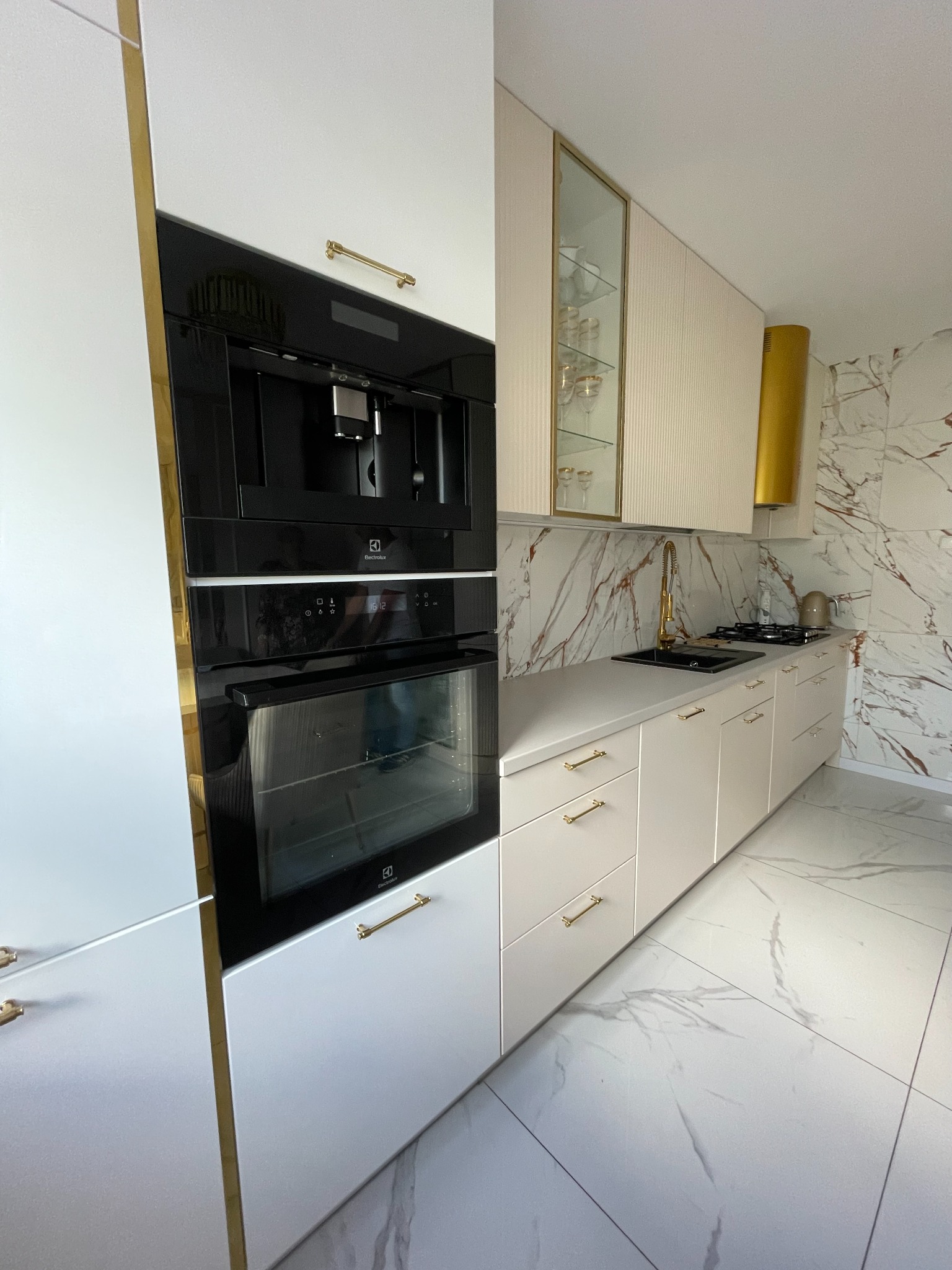 Nowoczesna, jasna kuchnia z marmurowym wzorem na ścianie i podłodze. Zabudowa kuchenna z białymi frontami, złotymi uchwytami i ramami, wbudowany ekspres do kawy i piekarnik Electrolux.