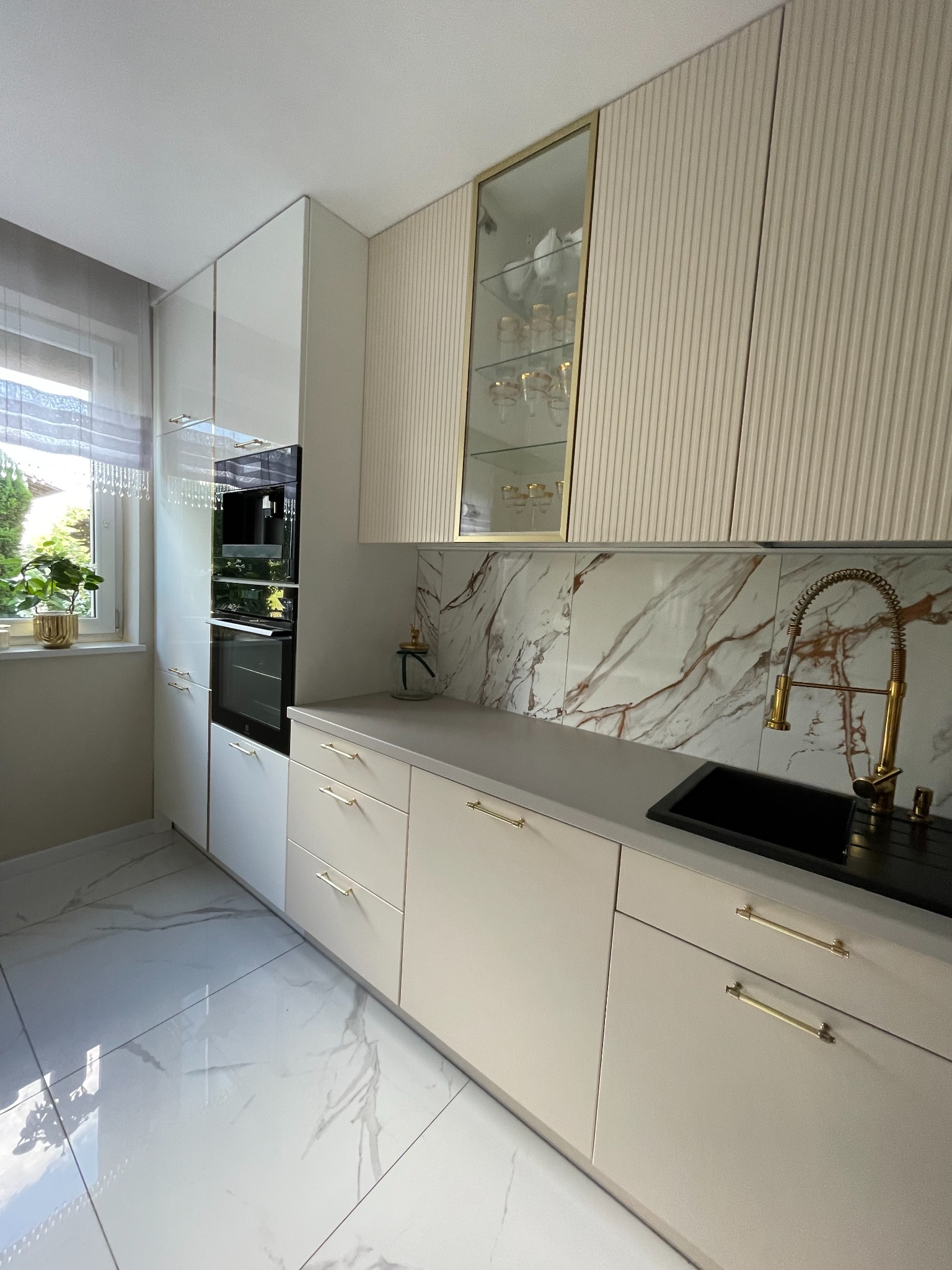 Nowoczesna kuchnia z białymi, lakierowanymi frontami, złotymi uchwytami i armaturą, marmurowym backsplash'em oraz podłogą z płytek imitujących marmur.