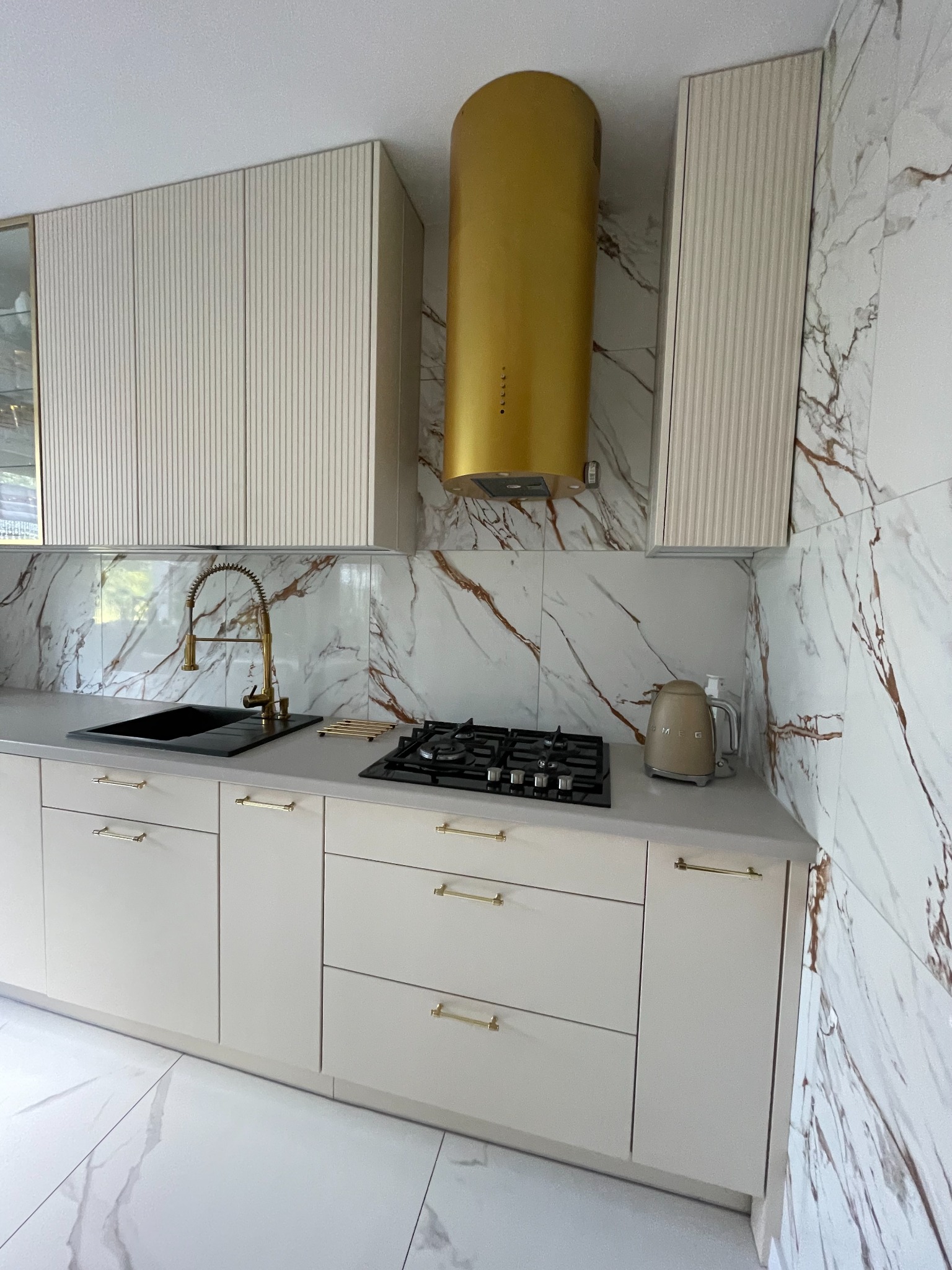 Nowoczesna kuchnia z beżowymi frontami szafek z delikatnym frezowaniem, złotymi uchwytami i złotym okapem na tle marmurowych płytek.