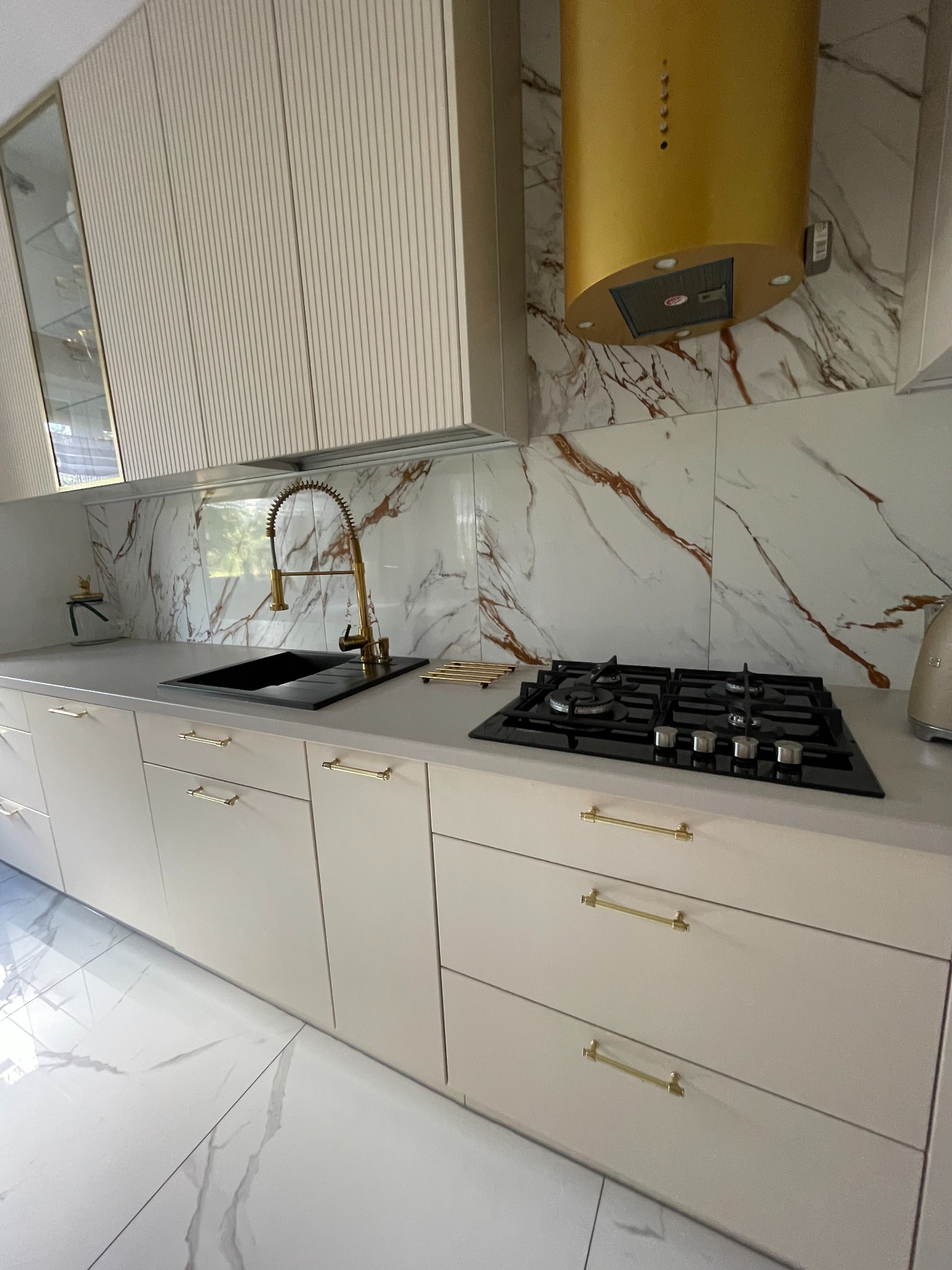 Nowoczesna kuchnia z beżowymi frontami szafek, złotymi uchwytami, marmurowym blatem i złotą armaturą oraz okapem.