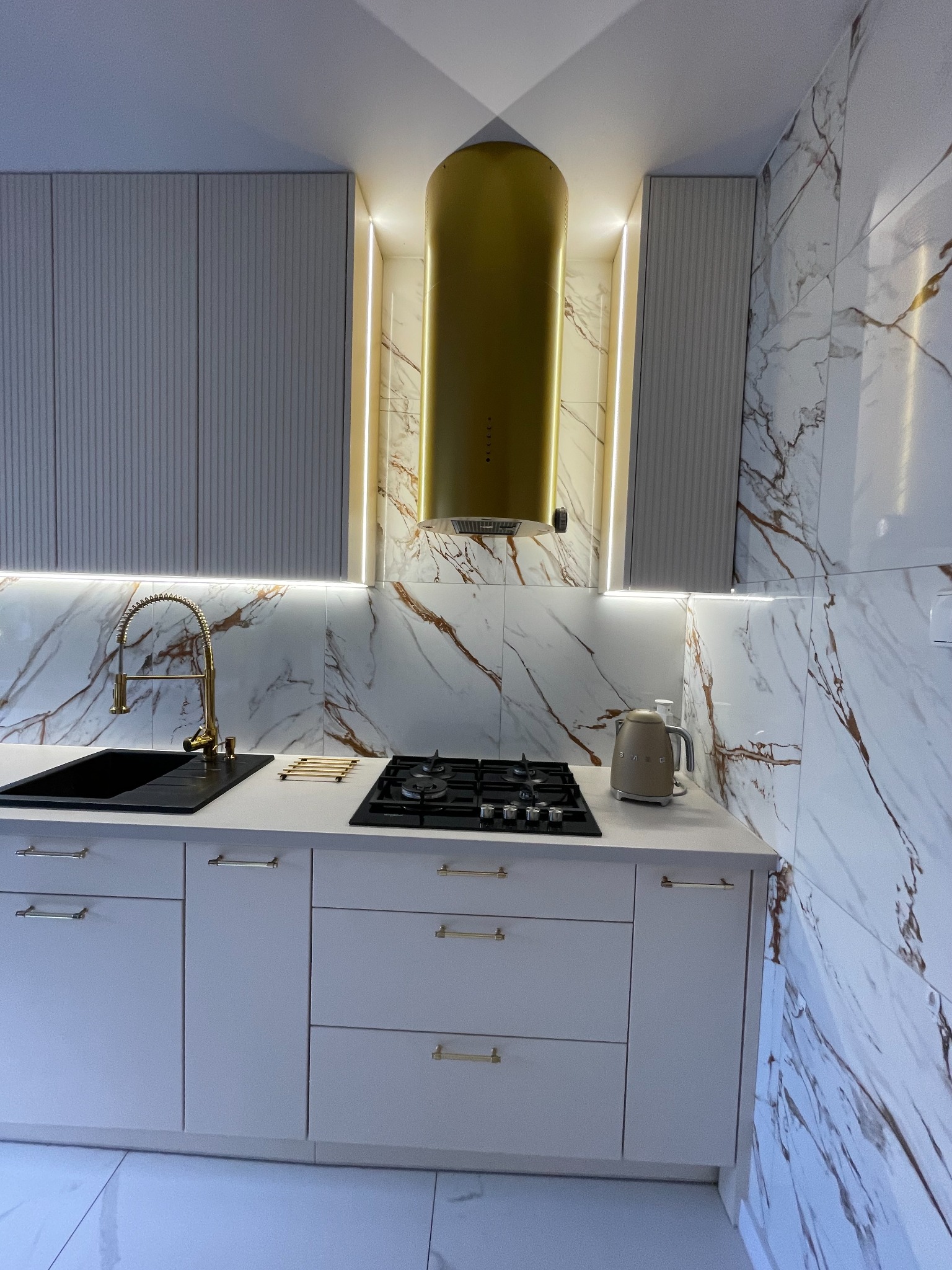 Nowoczesna kuchnia z białymi frontami szafek, złotymi akcentami w postaci baterii i uchwytów, marmurowymi płytkami na ścianie oraz złotym okapem cylindrycznym, oświetlona taśmami LED.