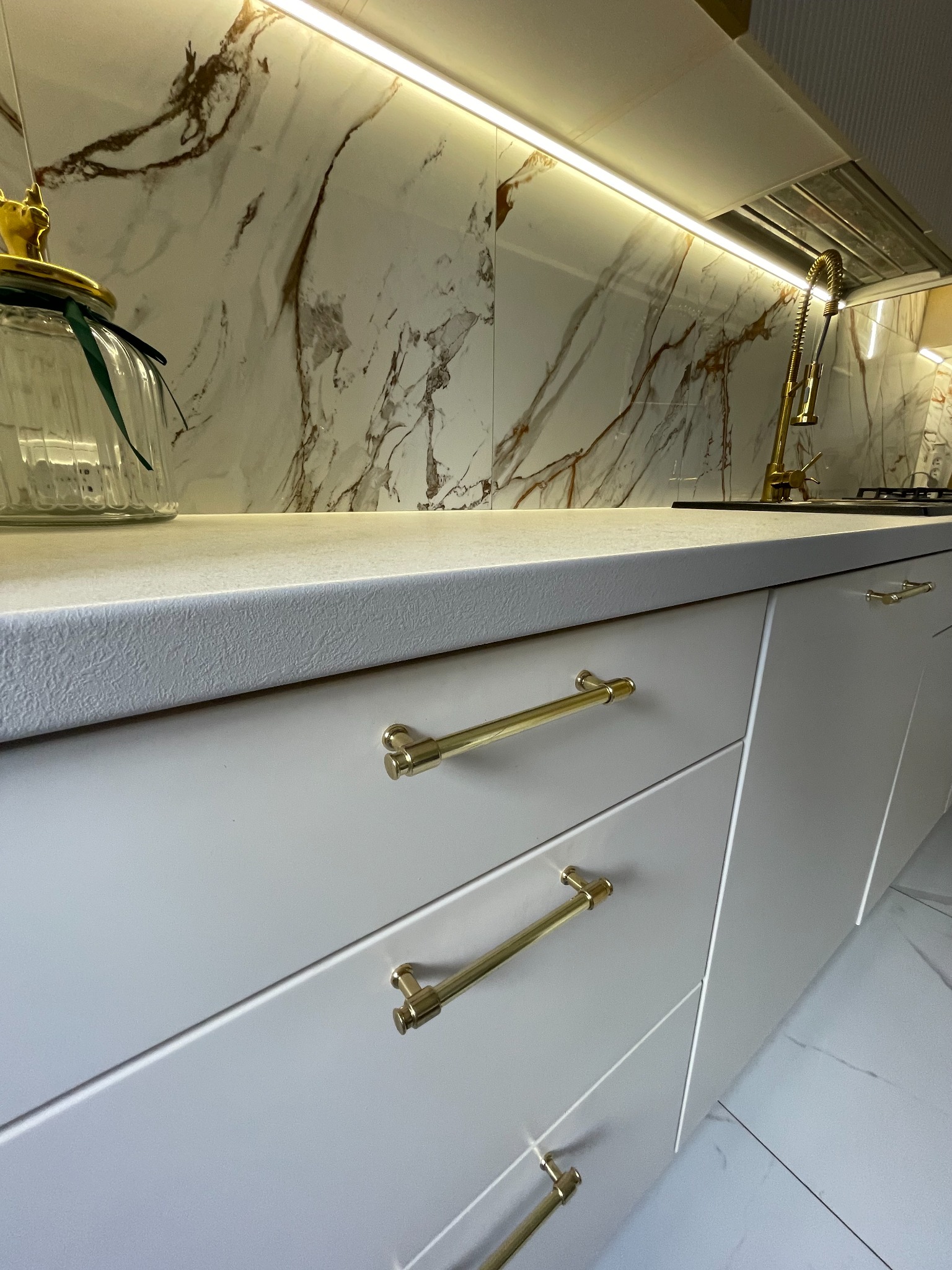 Szare fronty szafek kuchennych z złotymi uchwytami, kremowy blat z wyraźną fakturą, marmurowe płytki na ścianie podświetlone ledowym paskiem.