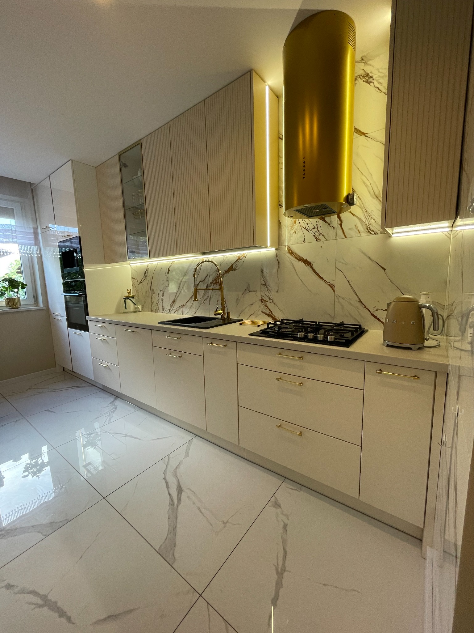 Jasna kuchnia z marmurowym backsplashiem, złotymi akcentami i podświetleniem LED, widoczny złoty okap cylindryczny, czarna płyta gazowa i beżowy czajnik Smeg.