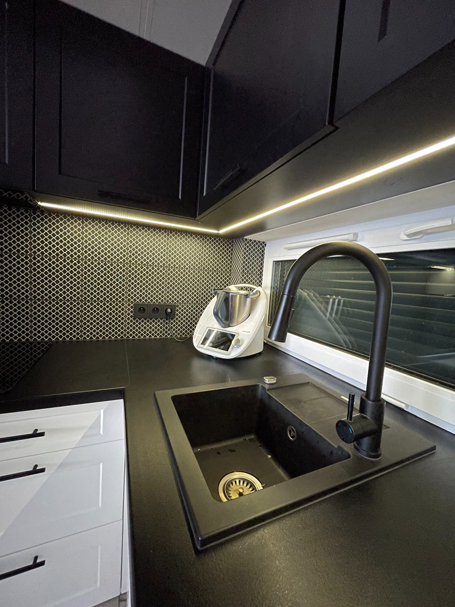 Czarna kuchnia z białymi szafkami dolnymi, czarnym blatem i zlewozmywakiem, oświetleniem LED pod szafkami wiszącymi i mozaiką na ścianie.