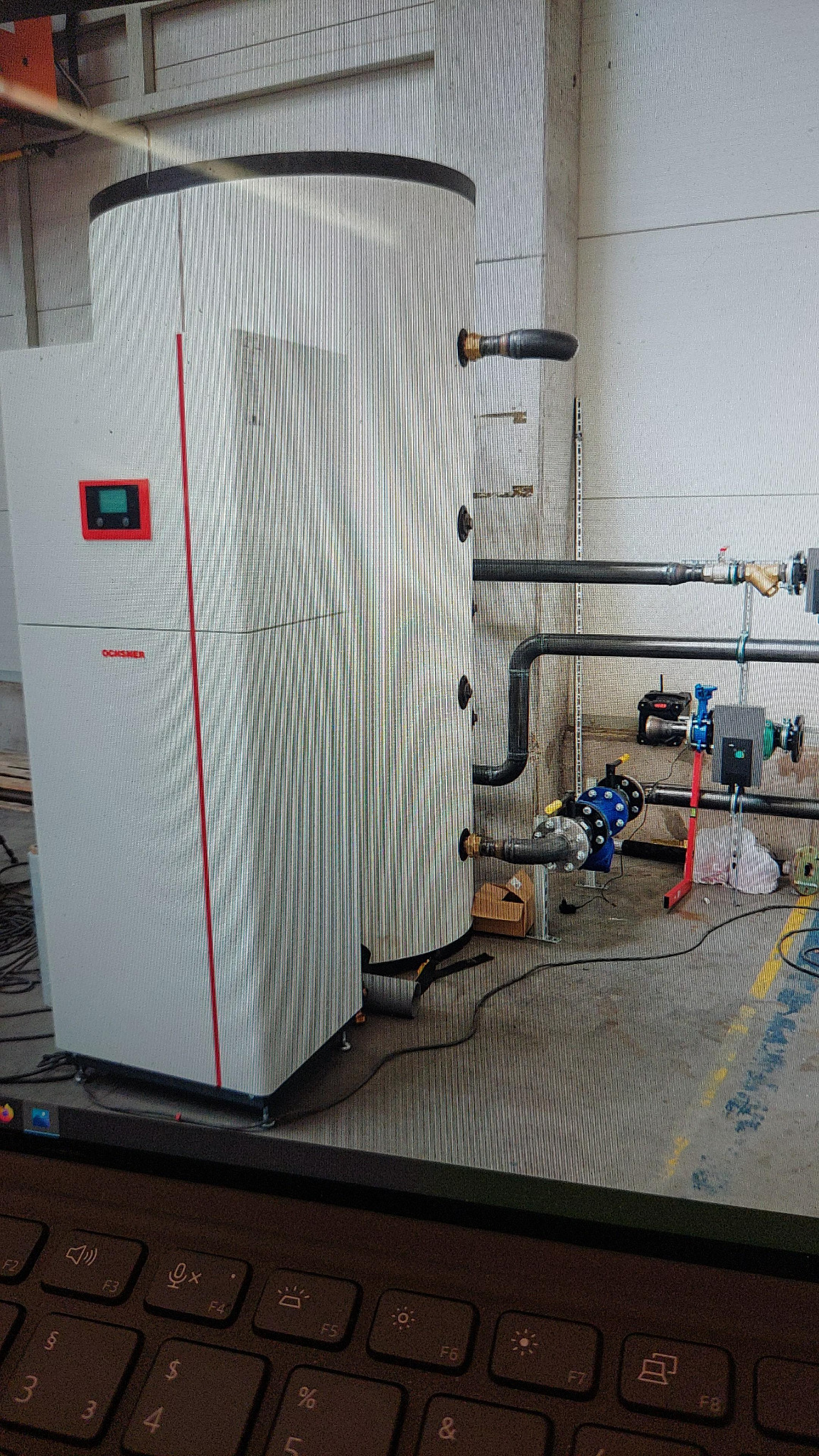 Biały moduł z czerwonym akcentem i wyświetlaczem, prawdopodobnie element instalacji grzewczej, podłączony grubymi rurami do instalacji w pomieszczeniu technicznym.